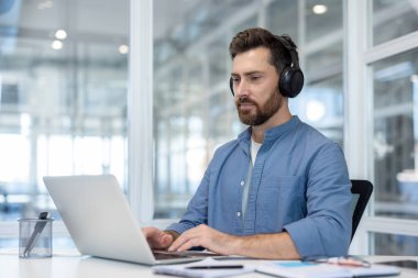 Man with beard and headphones concentrating on work, typing on a laptop computer while sitting at a desk in a contemporary, light-filled office environment