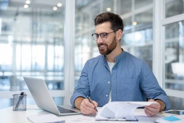 Young businessman working confidently at his desk, reviewing documents and using a laptop with a positive expression, symbolizing productivity and career success in a modern workplace