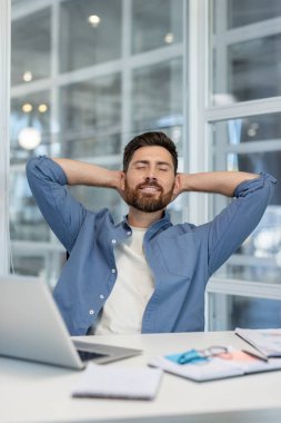 Smiling man finding serenity at his office desk, leaning back with hands behind his head, enjoying a moment of relaxation and satisfaction after achieving a task with his laptop nearby