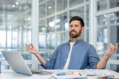 Businessman performing a meditation pose with hands in jnana mudra, finding calm, reducing stress, and achieving work-life balance inside a modern office workspace