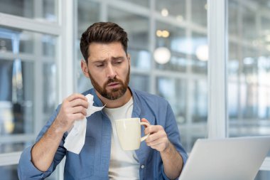Unwell man experiencing cold symptoms, holding a tissue and a mug while working on his laptop, highlighting the challenge of illness impacting productivity in a home office setting
