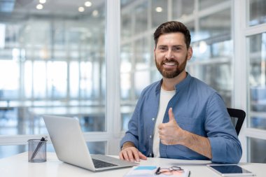 Smiling young professional at a modern desk giving a thumbs up to camera, confident and satisfied while working on his laptop, conveying approval, success and positivity