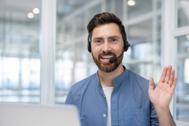 Man with beard and headset smiling, waving hand while making a video call, representing online customer support, virtual communication, and remote work from a modern office environment