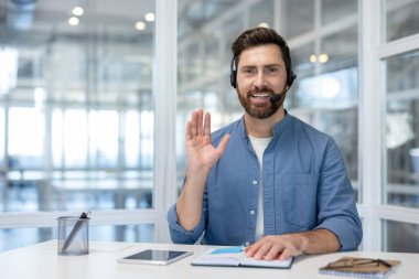 Friendly male call center agent wearing headset waving hand while smiling, participating in an online video conference from a modern office workspace, providing customer service and remote support