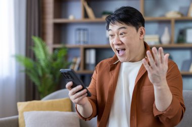 Surprised asian man receiving good news on smartphone, celebrating a win or success while sitting relaxed on a couch at home, showing extreme happiness and joy