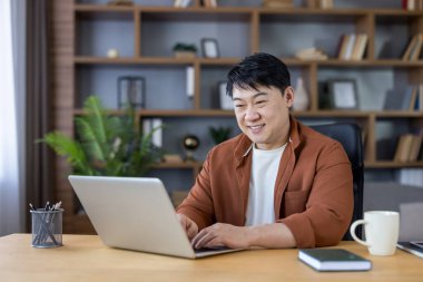 Asian man smiling while working remotely from a modern home office, typing on a laptop with shelves of books and a plant in the background, enjoying a comfortable and flexible work environment