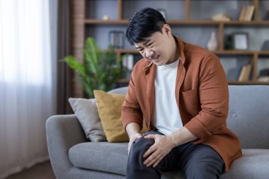 Middle-aged asian man sitting on sofa at home, grimacing and holding his knee in pain from injury or arthritis, showing discomfort and joint inflammation indoors