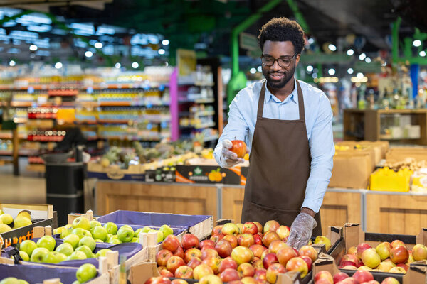 Male shop assistant wearing an apron and gloves, standing in a supermarket produce aisle and happily holding a fresh red apple, ensuring quality for customers
