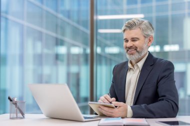 Mature businessman working at his desk in a modern office, happily writing notes in a notebook while looking at a laptop computer, representing productivity and professional engagement