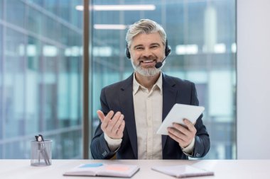 Mature man with a headset smiling and holding a digital tablet, representing concepts of online consultation, customer support, and virtual communication from a modern office environment