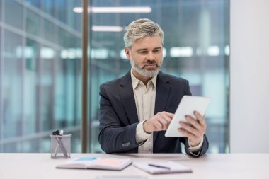 Businessman with a beard using a digital tablet at a modern office desk, interacting with technology while focusing on work tasks and corporate communication