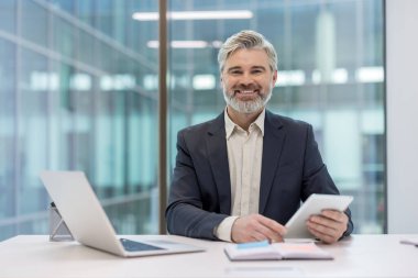 Experienced businessman sitting at a modern office desk, smiling at camera while holding a tablet, with laptop and planner nearby, conveying confidence, success and professionalism