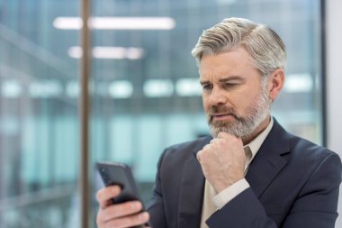 Mature corporate executive in a suit intently using a smartphone in a modern office, thinking and planning business strategy while checking messages and data for solutions