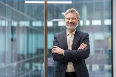 Mature businessman with a gray beard and professional attire, standing in a contemporary office setting with arms crossed and a confident, friendly smile