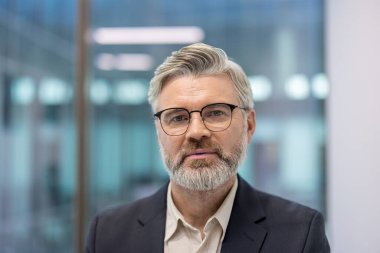 Senior businessman standing confidently in a modern office environment, wearing glasses and a suit, conveying professionalism, experience, and leadership in a corporate setting