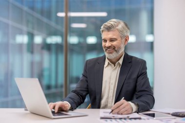 Mature businessman working at office desk, typing on laptop keyboard and making notes on a document with a pen, looking focused and smiling pleasantly