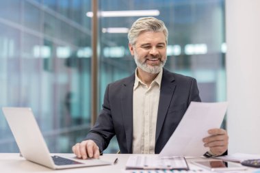 Mature gray-haired businessman with beard working at a modern desk, smiling while using a laptop and reviewing financial documents and reports, confident and productive in office