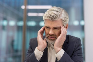 Senior businessman with gray hair and a beard, feeling intense pain and headache, holding temples with both hands, showing expressions of stress, frustration, and worry