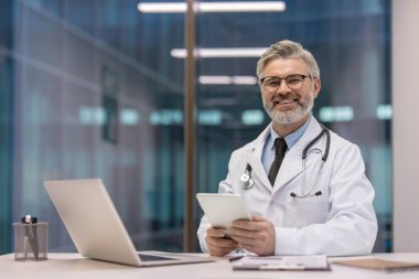 Experienced male doctor smiling and holding a digital tablet, sitting at a desk with a laptop, reflecting modern healthcare technology in a professional clinic setting
