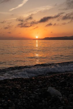 Kumsalda gün batımı dağın yanındaki suya düşüyor.