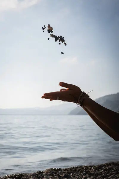Arka planda mavi deniz varken eller havaya taş atıyor.