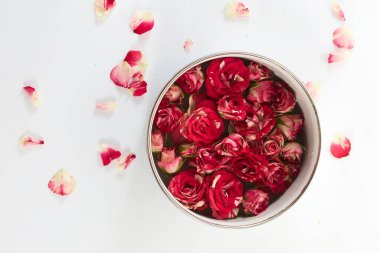 Su ve kırmızı güllerle dolu bir kase, beyaz bir yüzeyde dağınık yapraklarla çevrili..