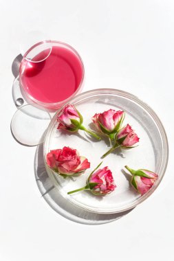 Pembe güller, pembe sıvı, beyaz zemin zeminli bir fincanın yanında berrak bir tabakta yatar..