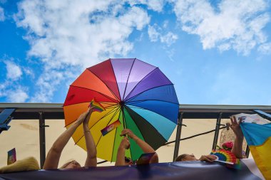Canlı bir gökkuşağı şemsiyesi kabarık bulutlu açık mavi bir gökyüzüne dayanıyor. Altındaki insanlar bayrak sallıyor..