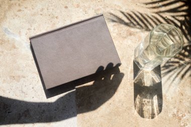 Bir ciltli kitap ve bir bardak su, güneşli bir masada gölgeler..