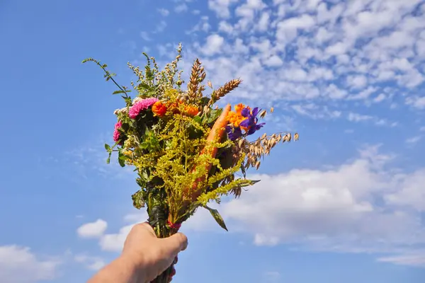 stock image A vibrant bouquet lifted against a partly cloudy blue sky, held by a hand. The colors are bright and warm.