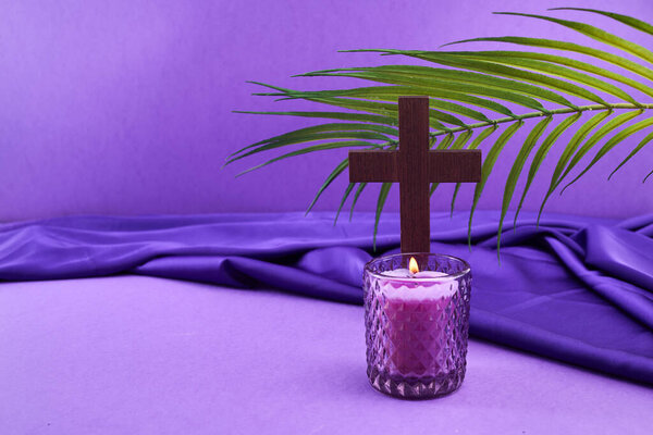 A wooden cross with a palm leaf and a lit purple candle on a purple fabric background.