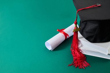 Mezuniyet şapkası, kırmızı püsküllü ve yeşil zemine karşı istiflenmiş kitaplar üzerine diploması var..
