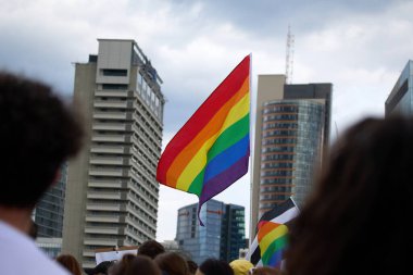Bir gurur bayrağı bulutlu bir gökyüzünün altında şehir ortamında kalabalığın arasında dalgalanıyor..