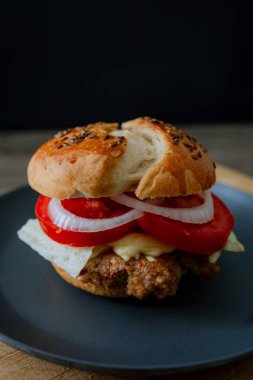 Siyah tabakta salatalık, domates ve marullu sığır burger, koyu besin estetiği ve karamsar arkaplan, dikey formatta yakalanır.