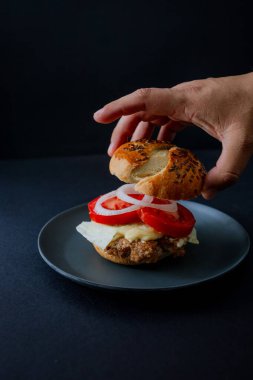 Siyah bir tabakta salatalık, domates ve marullu sığır burger, bir elle tutulan, kapkara bir yemek estetiği ve dengesiz bir arka planı var.