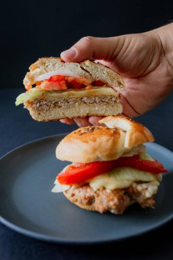 Salatalık dilimleri ve taze domatesli sulu bir sığır burger, siyah bir tabakta sunuluyor ve bir elle tutuluyor. Dramatik ışıklandırma ve karanlık arka plan yiyeceklerin dokusunu ve detaylarını vurguluyor, çarpıcı bir karanlık gıda estetiği elde ediyor.