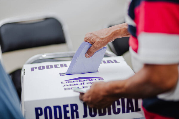 A hand places a ballot into a transparent box labeled Poder Judicial during Mexicos first-ever judicial election. The image captures citizen participation and electoral reform on a historic voting 