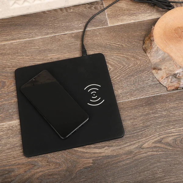 Charging the smartphone with leather wireless charger on desk. Black  leather charger pads. Wireless mousepad, Charger pad. Closeup, top view, no people. Concept shoot.
