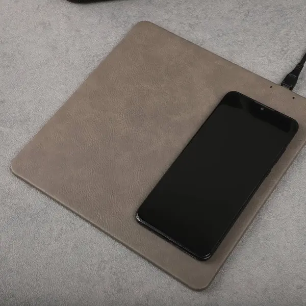 Charging the smartphone with leather wireless charger on desk. Gray leather charger pads. Wireless mousepad, Charger pad. Closeup, top view, no people. Concept shoot.