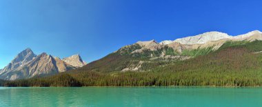Karla kaplanmış engebeli tepelerle dolu görkemli bir dağ sırasının nefes kesici manzarası canlı, mavi bir gökyüzüne karşı kurulmuş. Yeşillik yemyeşil orman, bu dağlık arazinin el değmemiş güzelliğini artırmak için üs boyunca uzanıyor..
