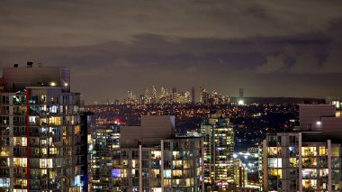 Geceleri modern bir şehir manzarasının nefes kesen panoramik görüntüsü, ön planda aydınlatılmış konutlar ve uzaklarda parlayan bir gökyüzü. Binaların yumuşak ışığı, karanlık ve kasvetli gökyüzü ile güzel bir tezat oluşturuyor..