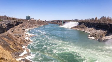 Niagara Şelalesi 'ndeki Amerikan Şelalesi' nin nefes kesici geniş açılı görüntüsü, arkada Kanada ve ABD 'yi birbirine bağlayan Gökkuşağı Köprüsü. Gözlem kulesi, şelalenin yanında yükselmekte. Güçlü çağlayandan yükselen sis gibi.. 