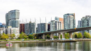 Vancouver şehir merkezinin modern deniz kenarı mimarisinin manzarası, sakin suların üzerine uzanan belirgin beton bir köprü. Görüntü, yüksek binalar ve ticari binalar ile küçük renkli bir su taksisinin karışımını yakalıyor.. 