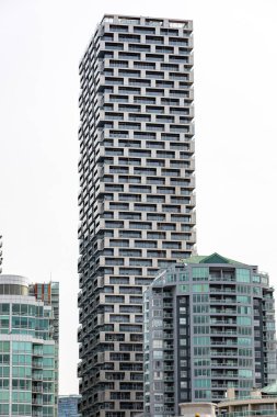 Şehir siluetindeki diğer modern binaların arasında dimdik duran, geometrik cepheli, çağdaş, yüksek katlı konut kulesinin görüntüsü. Görüntü, metropolde yaşayan canlıların temiz çizgilerini, mimari simetrisini ve modern tasarım unsurlarını yakalıyor..