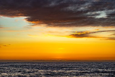 Bu çarpıcı deniz manzarası, kırmızı, turuncu ve mor tonlarda parlayan bir yaz günbatımının büyüsünü yakalar. Yumuşak okyanus dalgaları engebeli kayalık kıyılarla buluşup huzurlu ama dramatik bir kıyı manzarası yaratıyor..