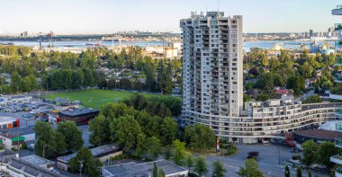 Bu büyüleyici çekim, yüksek kıvrımlı, yüksek eğimli bir bina, yemyeşil parklar ve uzaktaki vinçleri ve nakliye faaliyetleri ile doğa ve şehir yaşamını harmanlayan endüstriyel bir rıhtımın yer aldığı bir Vancouver mahallesinin panoramik görüntüsünü sunuyor..