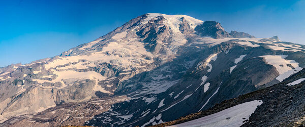 A stunning panoramic close-up view of a snow-capped mountain peak with glaciers, rugged rocky cliffs, and alpine slopes under a clear blue sky. Ideal for stock photography about wilderness, adventure, climbing, and nature exploration.