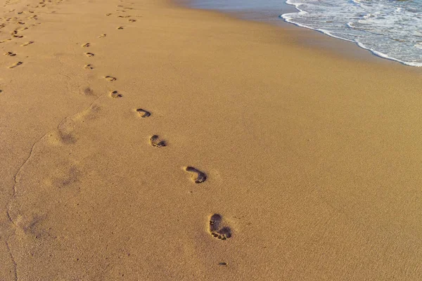 Human footprints on the yellow sand. Walking way near ocean water and foam in sun rays. Summer vacation on the coastline of a resort. Playa de Las Vistas, Tenerife, Spain
