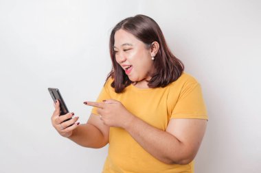 Asyalı iri yapılı mutlu bir kadının portresi gülümsüyor ve elinde akıllı telefonuyla beyaz bir arkaplan tarafından izole edilmiş sarı bir tişört giyiyor.