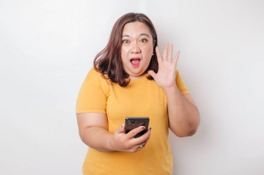 Sarı tişörtlü büyük Asyalı kadın akıllı telefonunu işaret ediyor, beyaz arka planla izole edilmiş.